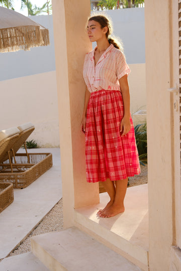 Introducing the Crimson Checkered Cuban Shirt, a collaboration between Audrey E Leary and World of Crow. This hand-woven jamdani fabric shirt is light pink with red motifs, short sleeves, and is super light and breathable, perfect for everyday wear. Sustainably made from soft cotton, paired with a red checkered skirt.