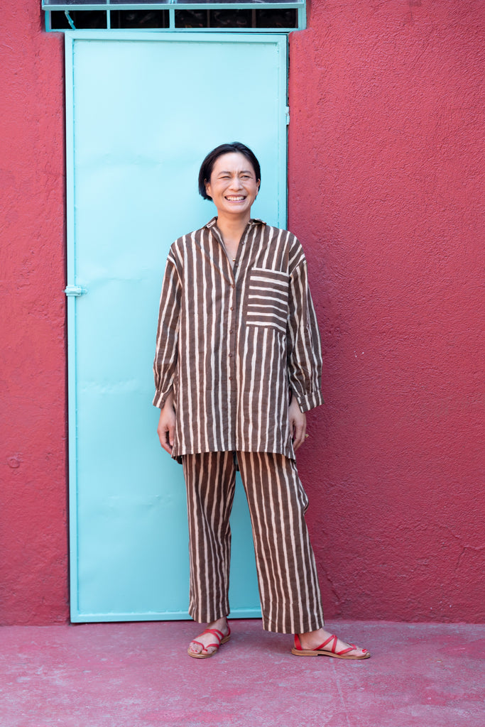 A brown and white striped long-sleeve shirt with a relaxed, oversized fit. The shirt features a classic pointed collar and a V-neck opening without buttons, giving it an effortless and casual look. A single chest pocket with horizontal stripes contrasts with the vertical pattern on the rest of the shirt, adding visual interest. The lightweight fabric drapes comfortably, making it ideal for a relaxed yet stylish outfit.