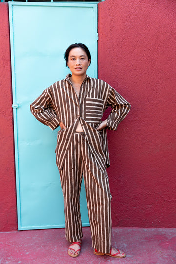 A brown and white striped long-sleeve shirt with a relaxed, oversized fit. The shirt features a classic pointed collar and a V-neck opening without buttons, giving it an effortless and casual look. A single chest pocket with horizontal stripes contrasts with the vertical pattern on the rest of the shirt, adding visual interest. The lightweight fabric drapes comfortably, making it ideal for a relaxed yet stylish outfit.