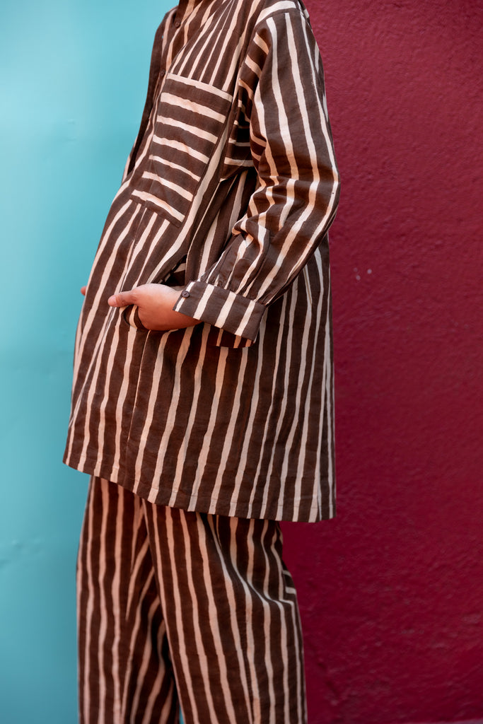 A brown and white striped long-sleeve shirt with a relaxed, oversized fit. The shirt features a classic pointed collar and a V-neck opening without buttons, giving it an effortless and casual look. A single chest pocket with horizontal stripes contrasts with the vertical pattern on the rest of the shirt, adding visual interest. The lightweight fabric drapes comfortably, making it ideal for a relaxed yet stylish outfit.