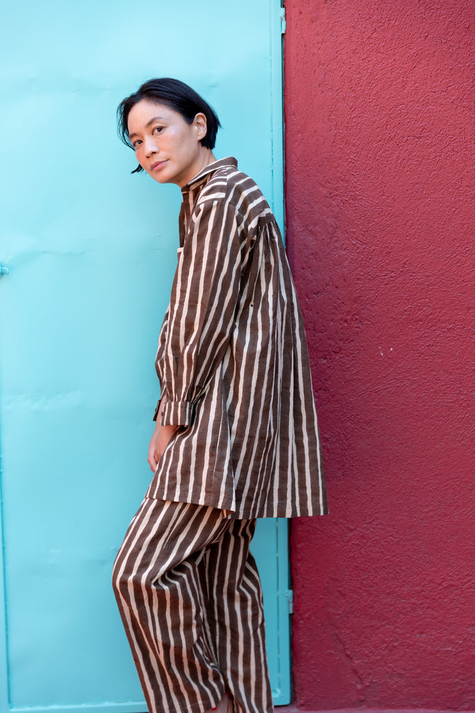 A brown and white striped long-sleeve shirt with a relaxed, oversized fit. The shirt features a classic pointed collar and a V-neck opening without buttons, giving it an effortless and casual look. A single chest pocket with horizontal stripes contrasts with the vertical pattern on the rest of the shirt, adding visual interest. The lightweight fabric drapes comfortably, making it ideal for a relaxed yet stylish outfit.