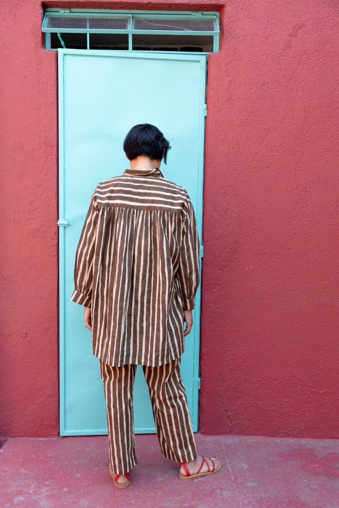 A brown and white striped long-sleeve shirt with a relaxed, oversized fit. The shirt features a classic pointed collar and a V-neck opening without buttons, giving it an effortless and casual look. A single chest pocket with horizontal stripes contrasts with the vertical pattern on the rest of the shirt, adding visual interest. The lightweight fabric drapes comfortably, making it ideal for a relaxed yet stylish outfit.