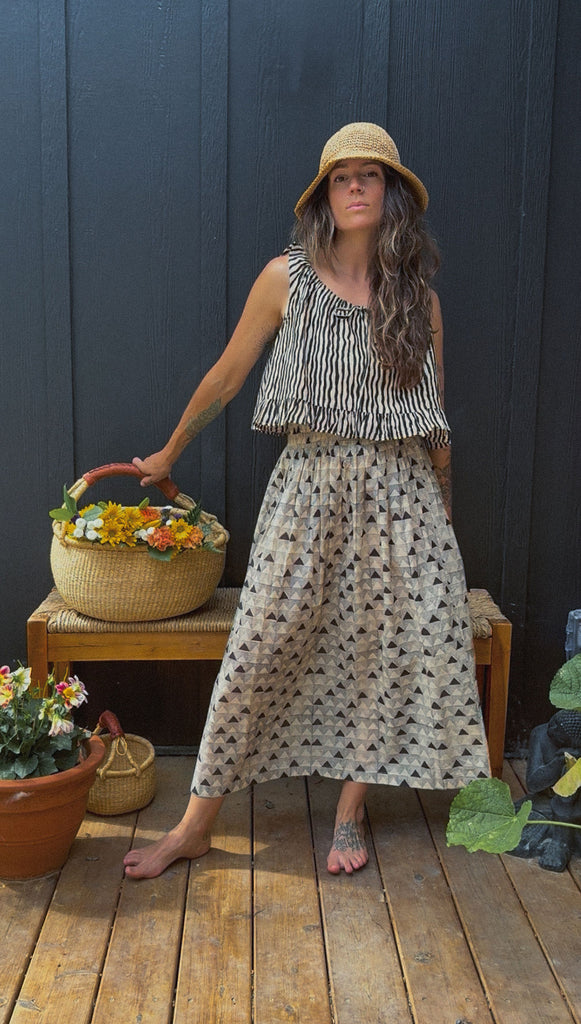 A mid-length, handwoven skirt made from soft, breathable organic cotton. The fabric features a delicate, all-over triangular block print in muted tones of black and grey on an off-white base. Lightly gathered at the waist, the skirt flows into a relaxed, voluminous silhouette that offers both comfort and movement. Naturally dyed and finished with a subtle texture, the skirt has an earthy, artisanal feel ideal for everyday wear.
