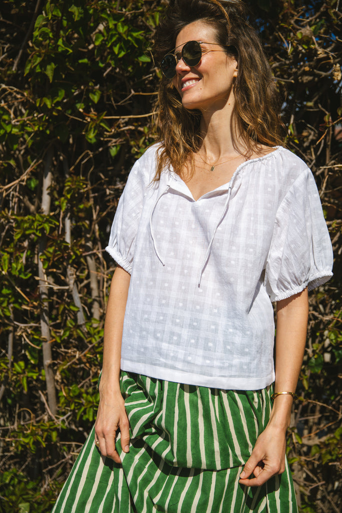 Lightweight white cotton top with a subtle textured weave, featuring a relaxed fit and airy design. The blouse has a gathered scoop neckline with delicate tie details, adding a touch of softness. Puff sleeves with gentle pleats create volume, while the slightly cropped hem gives it a modern, effortless feel.