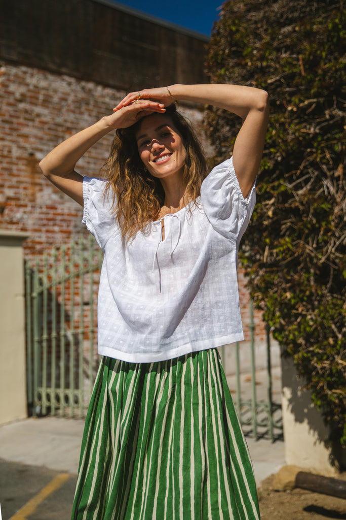 Lightweight white cotton top with a subtle textured weave, featuring a relaxed fit and airy design. The blouse has a gathered scoop neckline with delicate tie details, adding a touch of softness. Puff sleeves with gentle pleats create volume, while the slightly cropped hem gives it a modern, effortless feel.