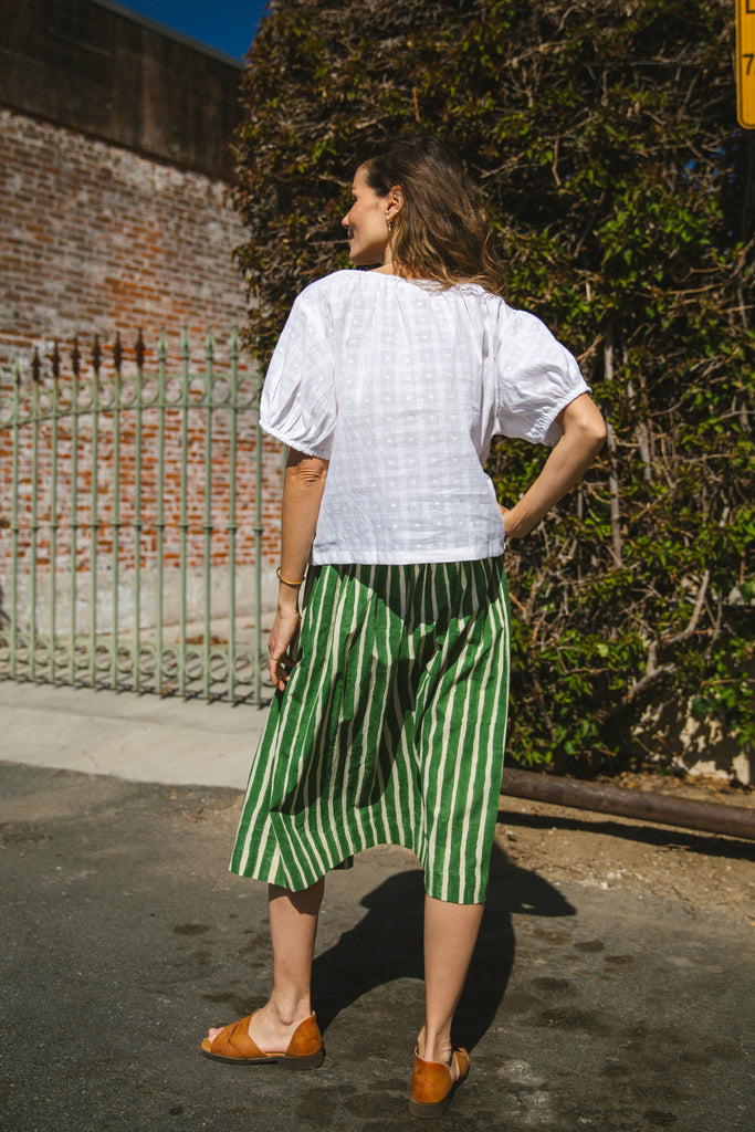 Lightweight white cotton top with a subtle textured weave, featuring a relaxed fit and airy design. The blouse has a gathered scoop neckline with delicate tie details, adding a touch of softness. Puff sleeves with gentle pleats create volume, while the slightly cropped hem gives it a modern, effortless feel.