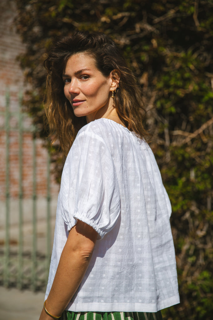 Lightweight white cotton top with a subtle textured weave, featuring a relaxed fit and airy design. The blouse has a gathered scoop neckline with delicate tie details, adding a touch of softness. Puff sleeves with gentle pleats create volume, while the slightly cropped hem gives it a modern, effortless feel.