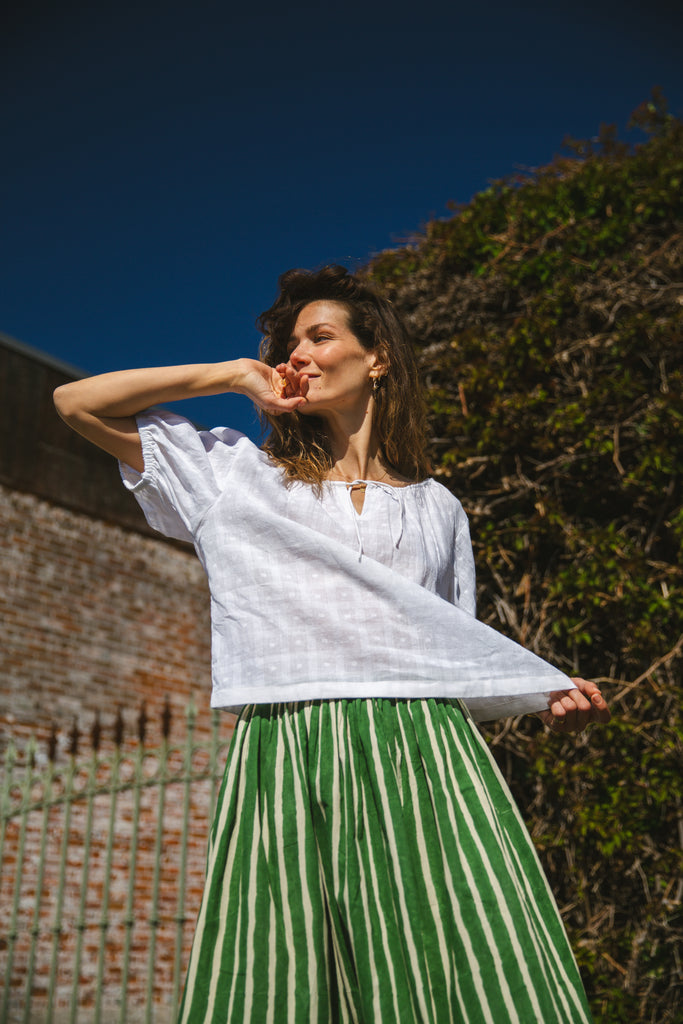 Lightweight white cotton top with a subtle textured weave, featuring a relaxed fit and airy design. The blouse has a gathered scoop neckline with delicate tie details, adding a touch of softness. Puff sleeves with gentle pleats create volume, while the slightly cropped hem gives it a modern, effortless feel.