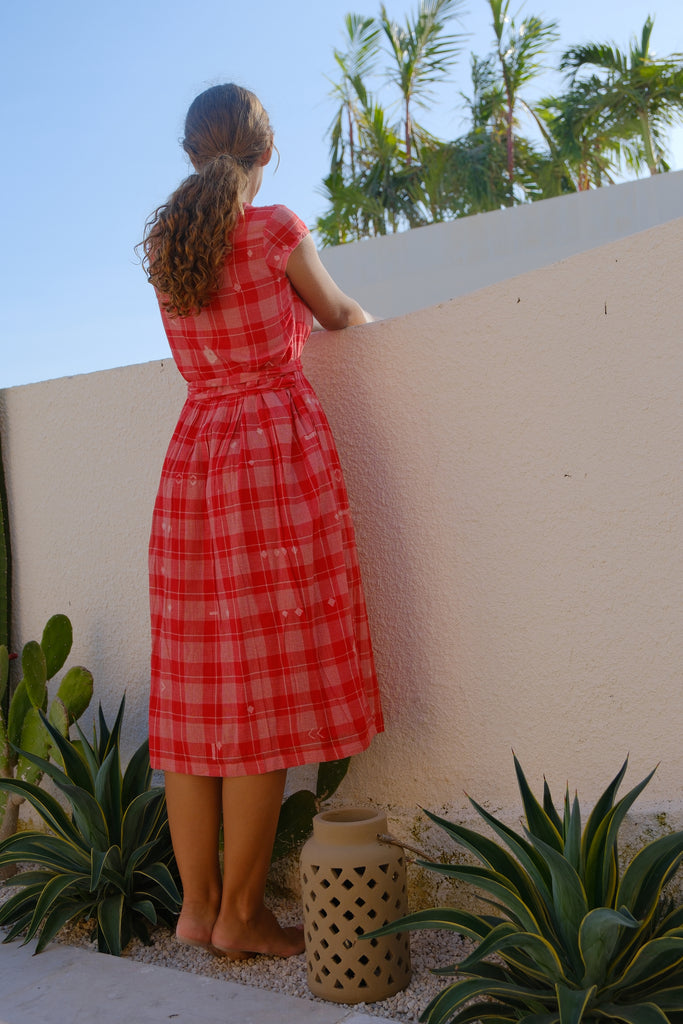 Introducing the Audrey Dress, a beautiful collaboration between Audrey E Leary and World of Crow. This wrap-on midi dress in a vibrant red color features a stylish V-neck and cap sleeves. Made from soft, handwoven cotton, it is sustainably crafted to offer both elegance and comfort. Seen on a woman, this dress provides a distinctive look with its unique design and sustainable materials.