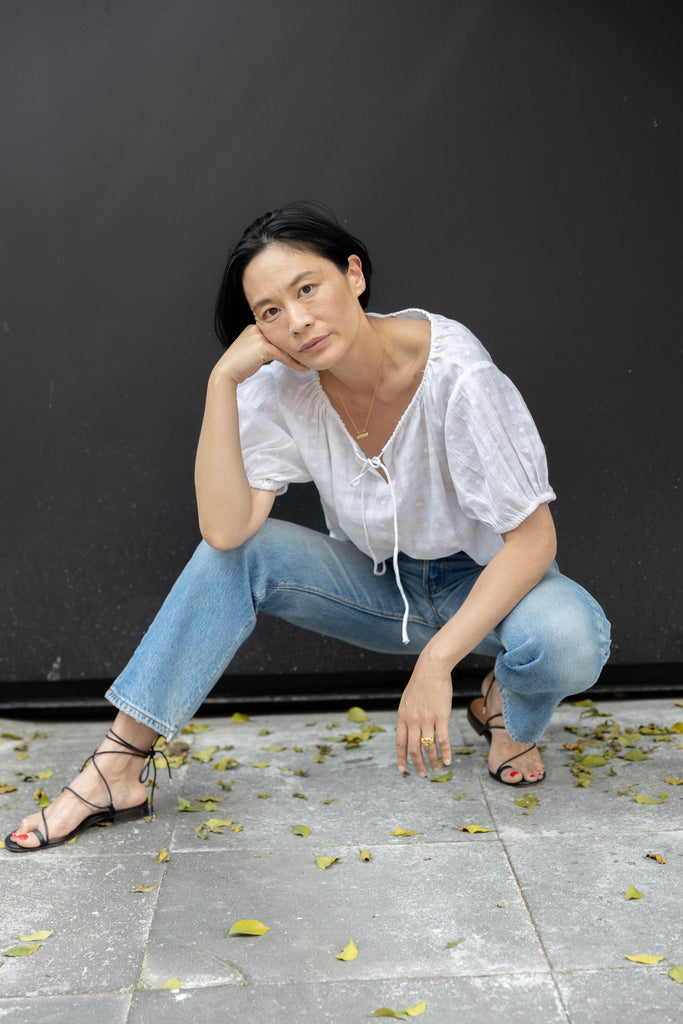 Lightweight white cotton top with a subtle textured weave, featuring a relaxed fit and airy design. The blouse has a gathered scoop neckline with delicate tie details, adding a touch of softness. Puff sleeves with gentle pleats create volume, while the slightly cropped hem gives it a modern, effortless feel.