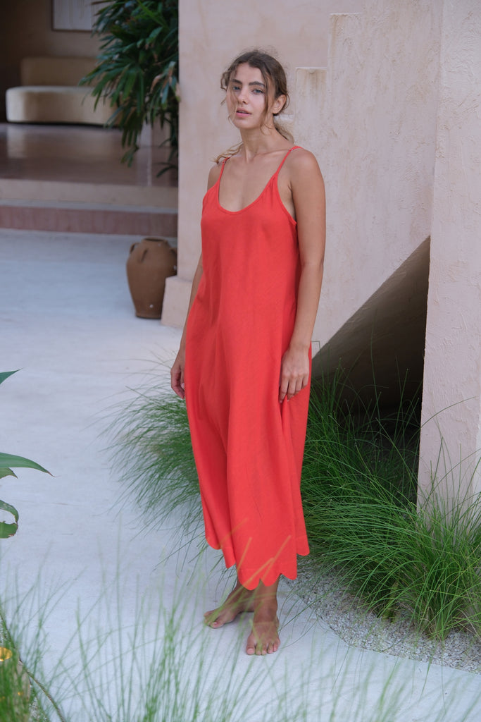 Introducing our Holiday Slip Dress, a collaboration between Audrey E Leary and World of Crow. This sleeveless dress in an orangish-red hue features scallop detailing and a midi length, crafted from handwoven cotton for a lightweight and breathable fit. Perfect for layering with a jacket, it's a versatile and sustainable choice for your outings