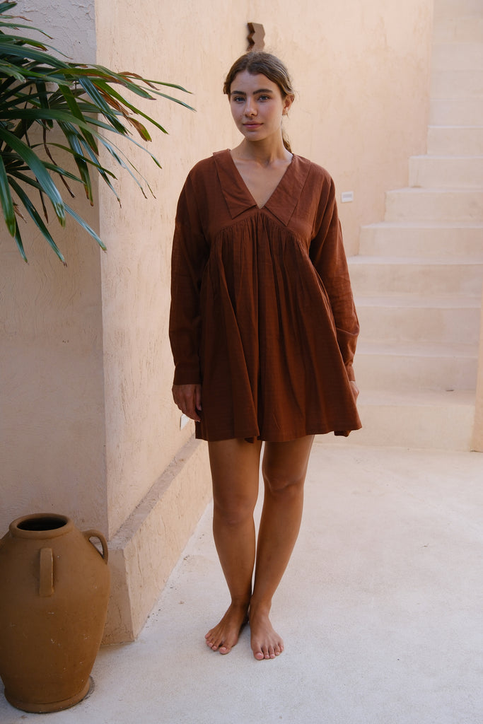 A woman stands on a patio in our Vintage Collar Dress, a collaboration between Audrey E Leary and World of Crow. This dress, in rich chocolate brown, features a classic collar and full sleeves, crafted from handwoven cotton for a refined and sustainable look
