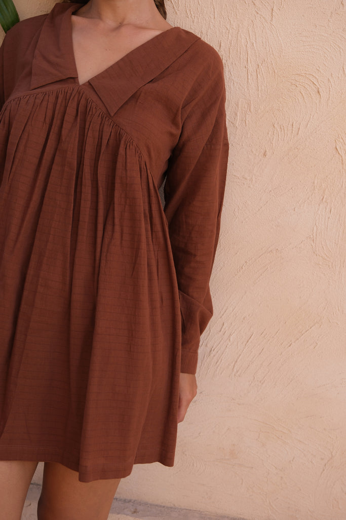 A woman stands on a patio in our Vintage Collar Dress, a collaboration between Audrey E Leary and World of Crow. This dress, in rich chocolate brown, features a classic collar and full sleeves, crafted from handwoven cotton for a refined and sustainable look