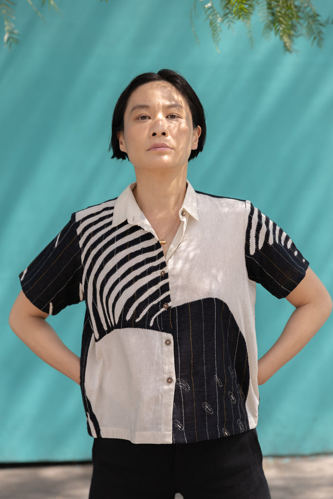 A short-sleeved, button-up shirt featuring a striking asymmetrical patchwork design in black, white, and beige. The fabric has a lightweight, textured feel, crafted from breathable natural fibers. The pattern blends bold geometric lines, organic curves, and subtle embroidered details, creating a dynamic contrast. The classic pointed collar adds a structured touch, while the relaxed, boxy fit gives it an effortlessly modern, artistic appeal.