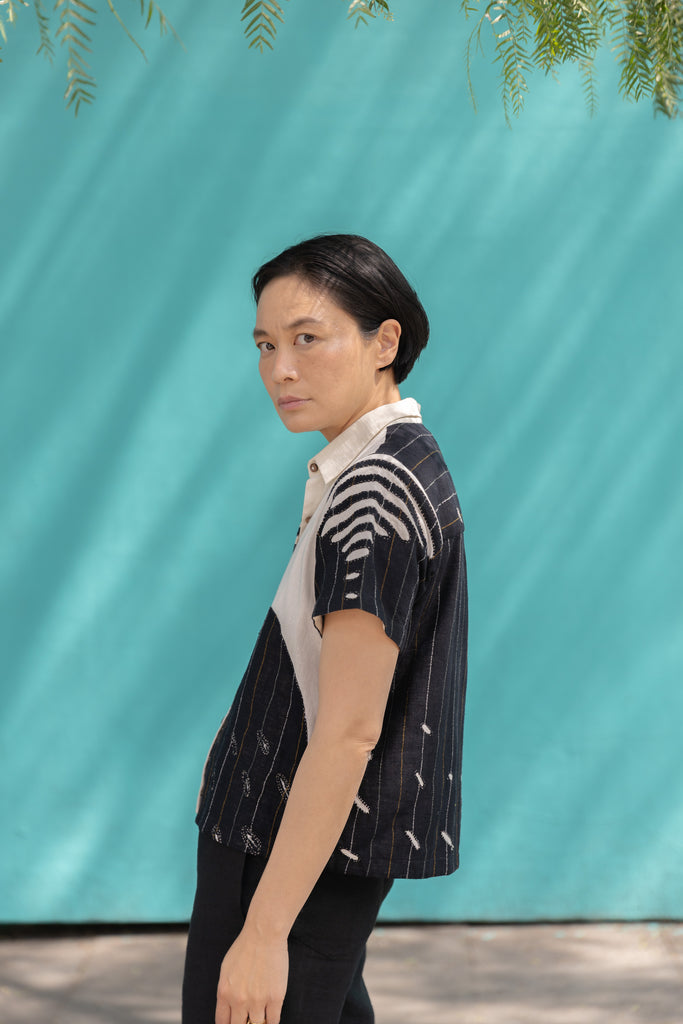 A short-sleeved, button-up shirt featuring a striking asymmetrical patchwork design in black, white, and beige. The fabric has a lightweight, textured feel, crafted from breathable natural fibers. The pattern blends bold geometric lines, organic curves, and subtle embroidered details, creating a dynamic contrast. The classic pointed collar adds a structured touch, while the relaxed, boxy fit gives it an effortlessly modern, artistic appeal.