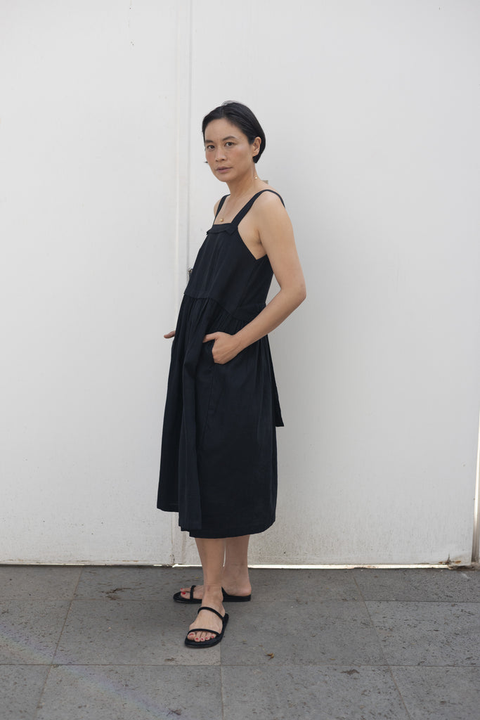 This is a black sleeveless midi dress with a structured square neckline and wide shoulder straps. The dress features a loose, flowing silhouette with a gathered waistline that adds volume and movement. Crafted from a lightweight, breathable fabric, it drapes effortlessly, making it perfect for warm weather. Functional side pockets enhance its practicality, while the relaxed fit exudes a laid-back yet refined charm. The minimalist design and deep black hue create a versatile piece that can be styled casually