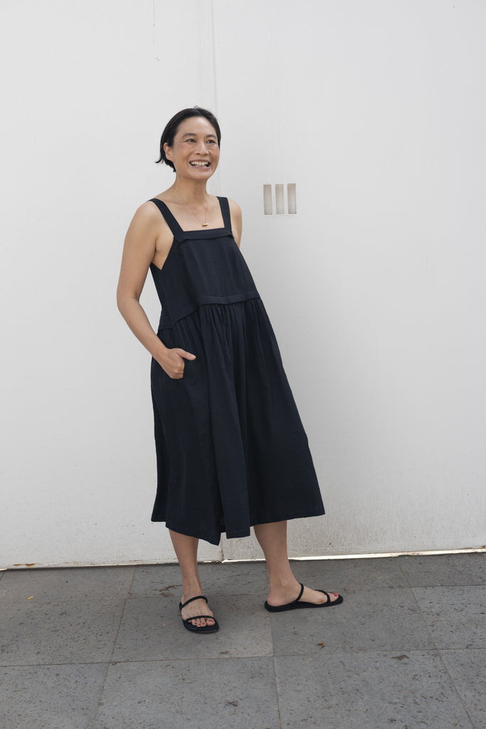 This is a black sleeveless midi dress with a structured square neckline and wide shoulder straps. The dress features a loose, flowing silhouette with a gathered waistline that adds volume and movement. Crafted from a lightweight, breathable fabric, it drapes effortlessly, making it perfect for warm weather. Functional side pockets enhance its practicality, while the relaxed fit exudes a laid-back yet refined charm. The minimalist design and deep black hue create a versatile piece that can be styled casually