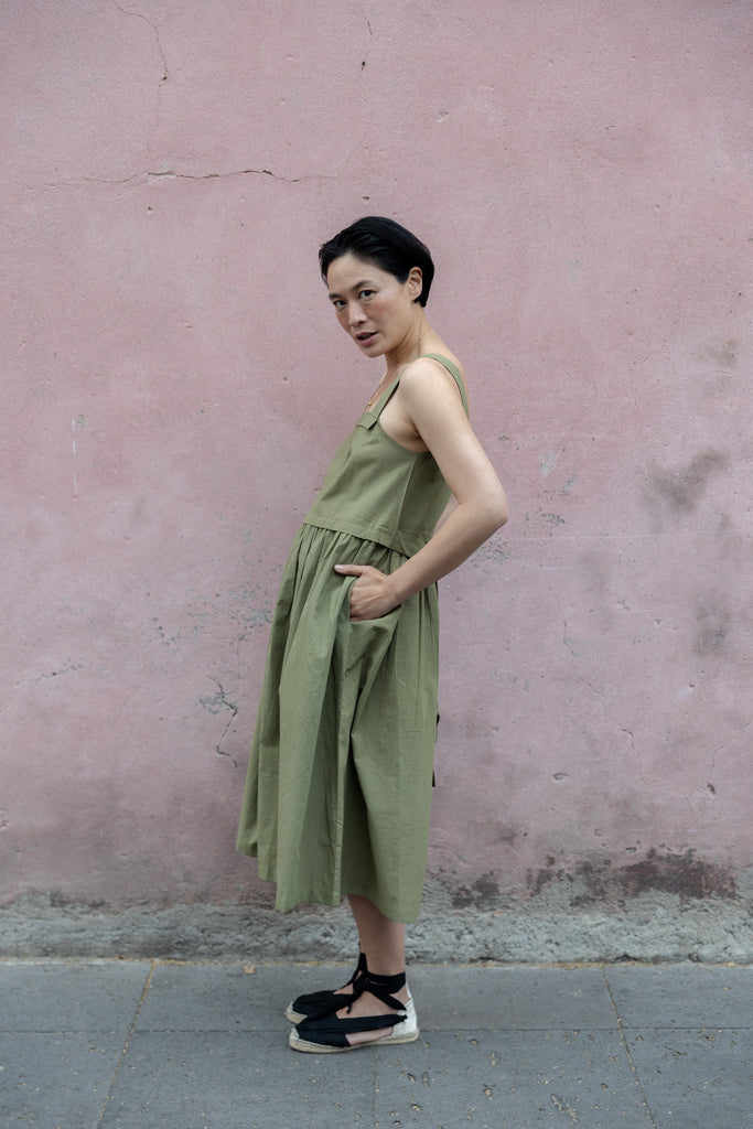 Olive green babydoll midi dress with wide shoulder straps. Features a gathered waistline and a voluminous, flowy skirt. Made from lightweight, structured fabric for a relaxed yet chic look.